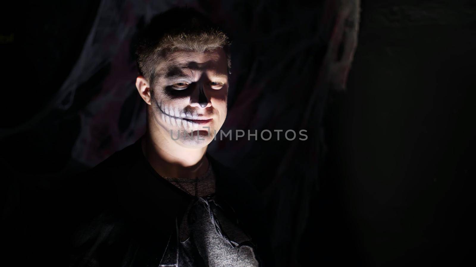 Halloween party, night, frightening portrait of a man in the twilight, in the rays of light. man with a terrible make-up in a black witch costume. High quality photo