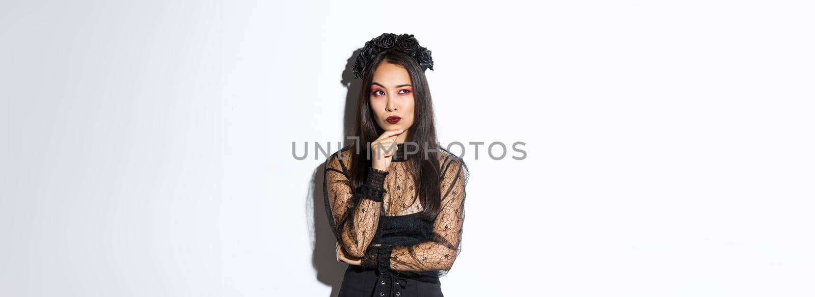 Thoughtful elegant evil witch in black lace dress, gothic wreath and makeup looking upper left corner. Woman making plans for halloween party, thinking over white background.