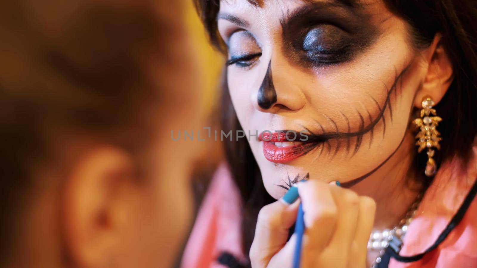 Halloween party, make-up artist draws a terrible makeup on the face of a brunette woman for a Halloween party. in the background, the scenery in the style of Halloween is seen. High quality photo