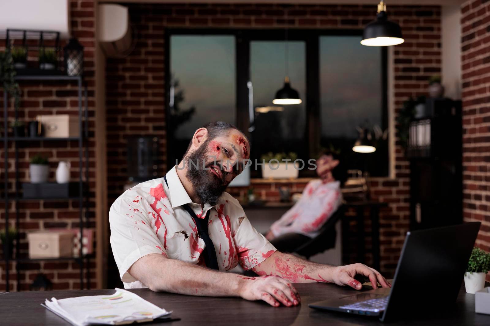 Portrait of zombie businessman sitting at desk, looking creepy and terrifiyng in company office. Brain eating cruel monster with bloody wounds working on laptop, horrible possessed devil.