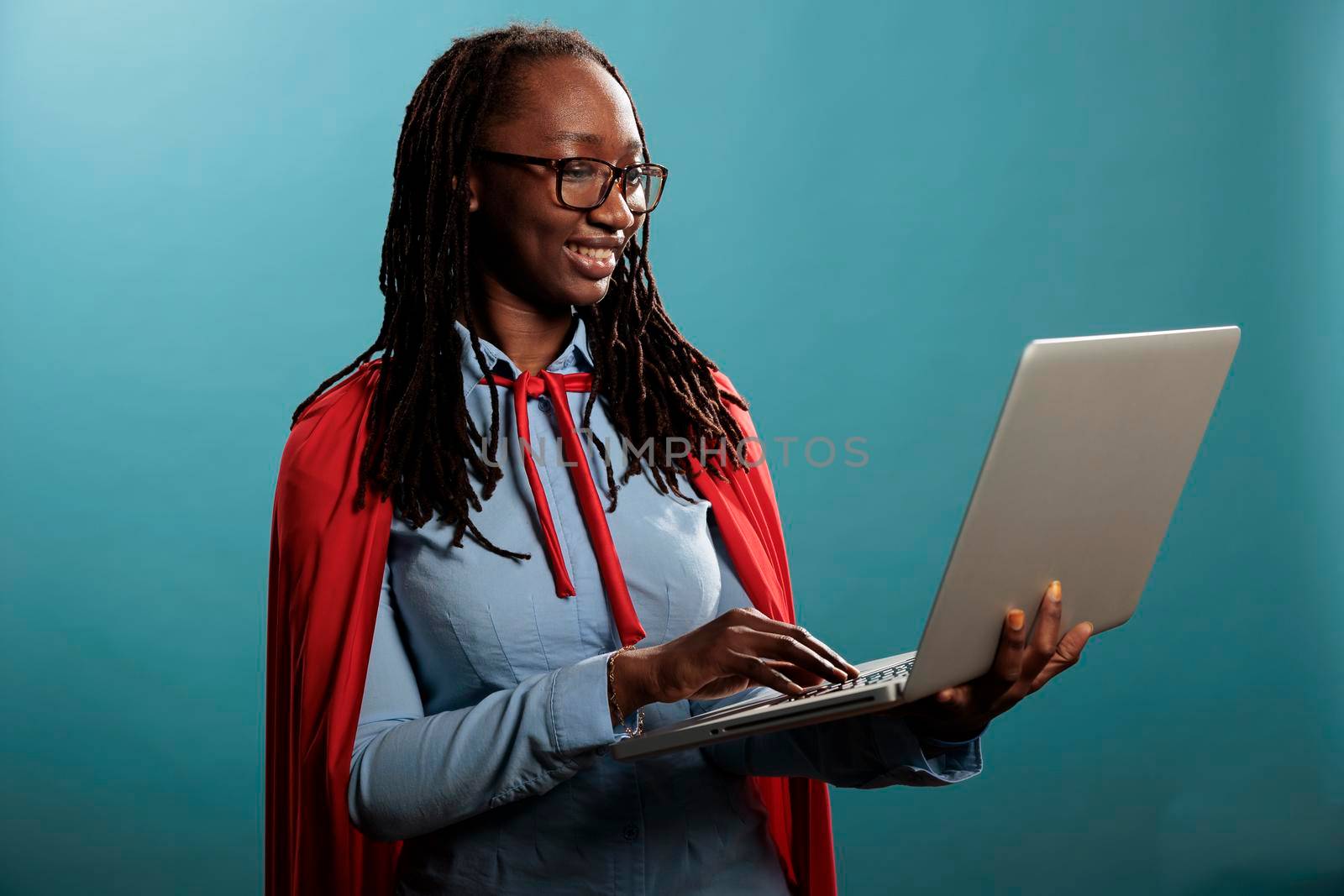 Portrait of confident and positive african american justice defender browsing internet on modern laptop. Happy joyful superhero woman wearing mighty hero red cloak while standing on blue background.