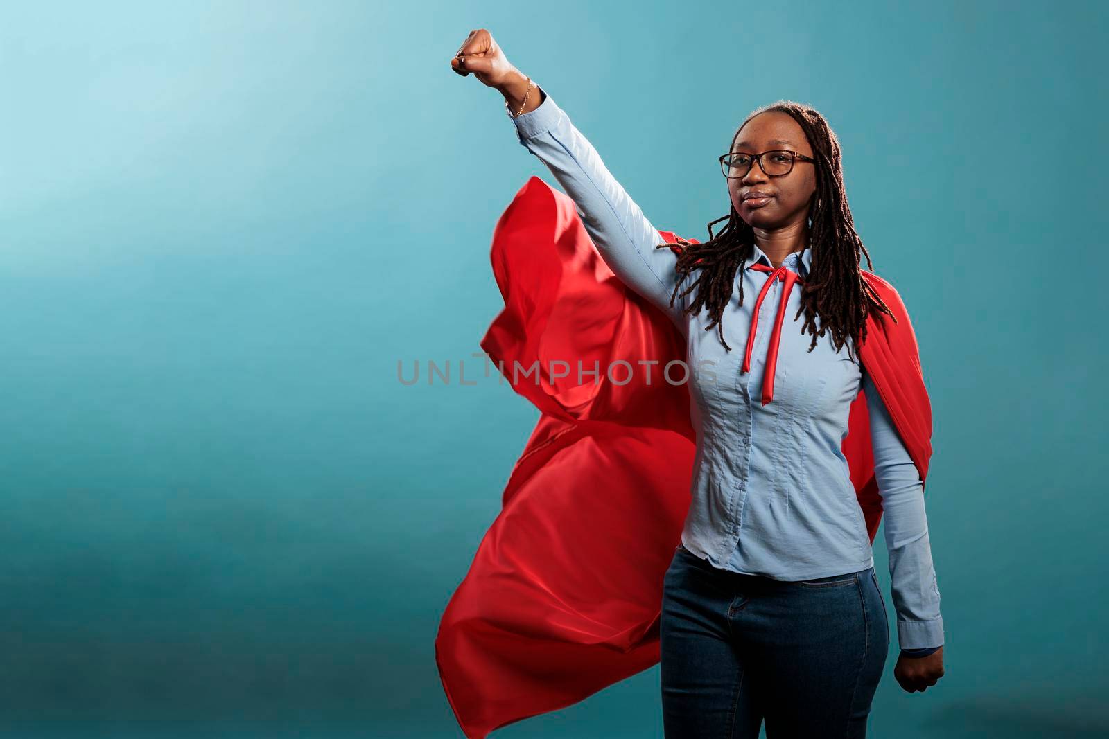 Brave and proud looking young adult superhero woman acting like a flying hero while wearing mighty cape on blue background. Strong justice defender with superpowers looking at camera with fist in air.
