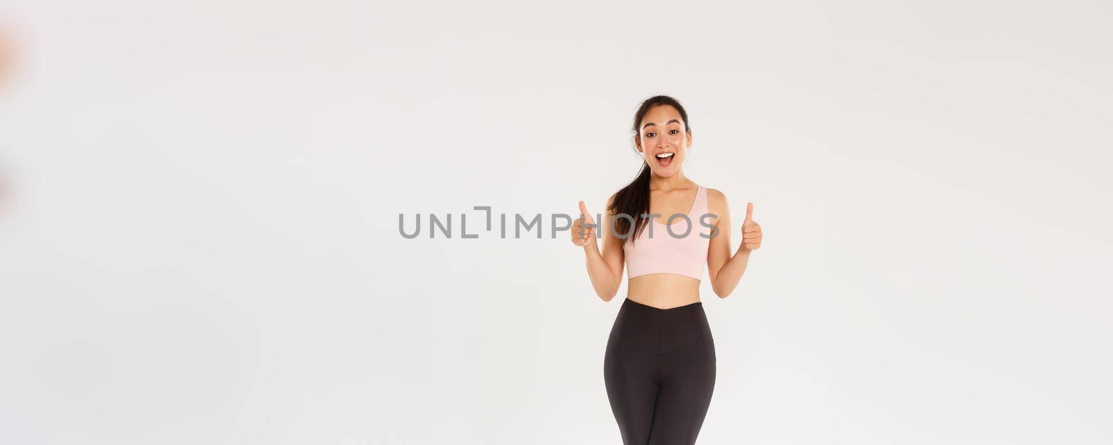 Full length of amazed and satisfied smiling fitness girl, female athelte in active wear liking new gym or workout program, showing thumbs-up pleased, encourage start training or doing exercises by Benzoix