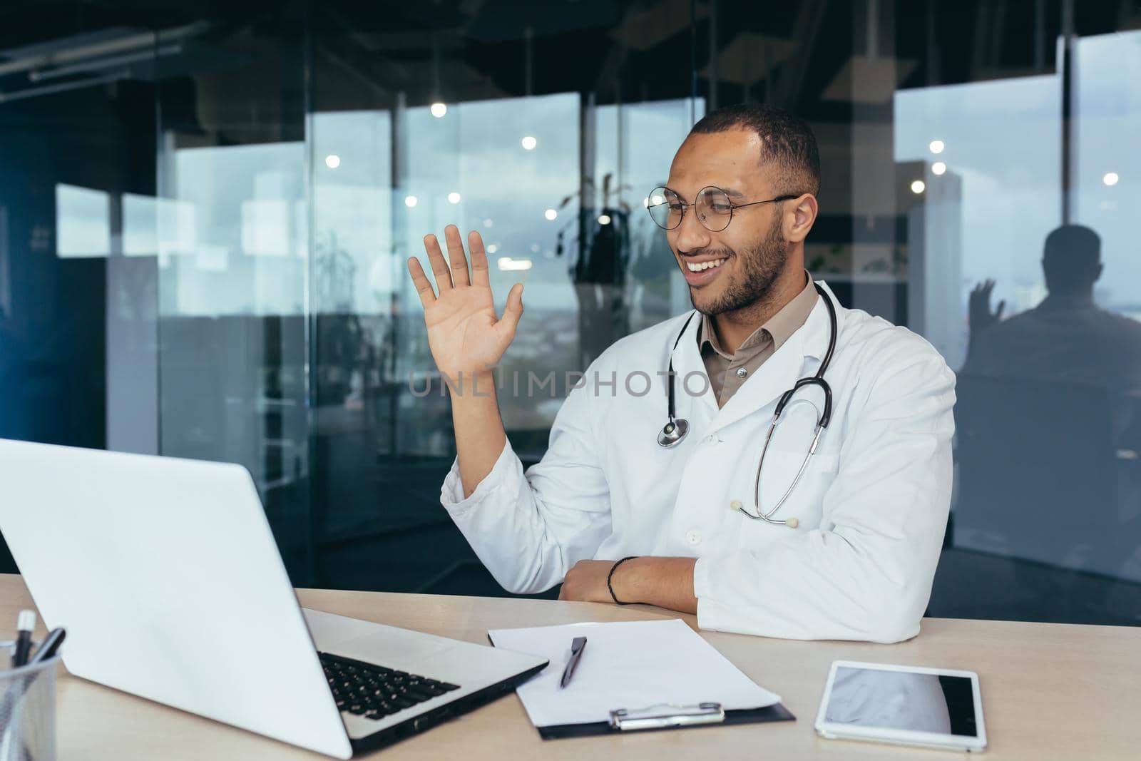 Happy hispanic doctor smiling and talking on video call man in white coat and stethoscope using laptop for remote online consultation smiling and waving hello.