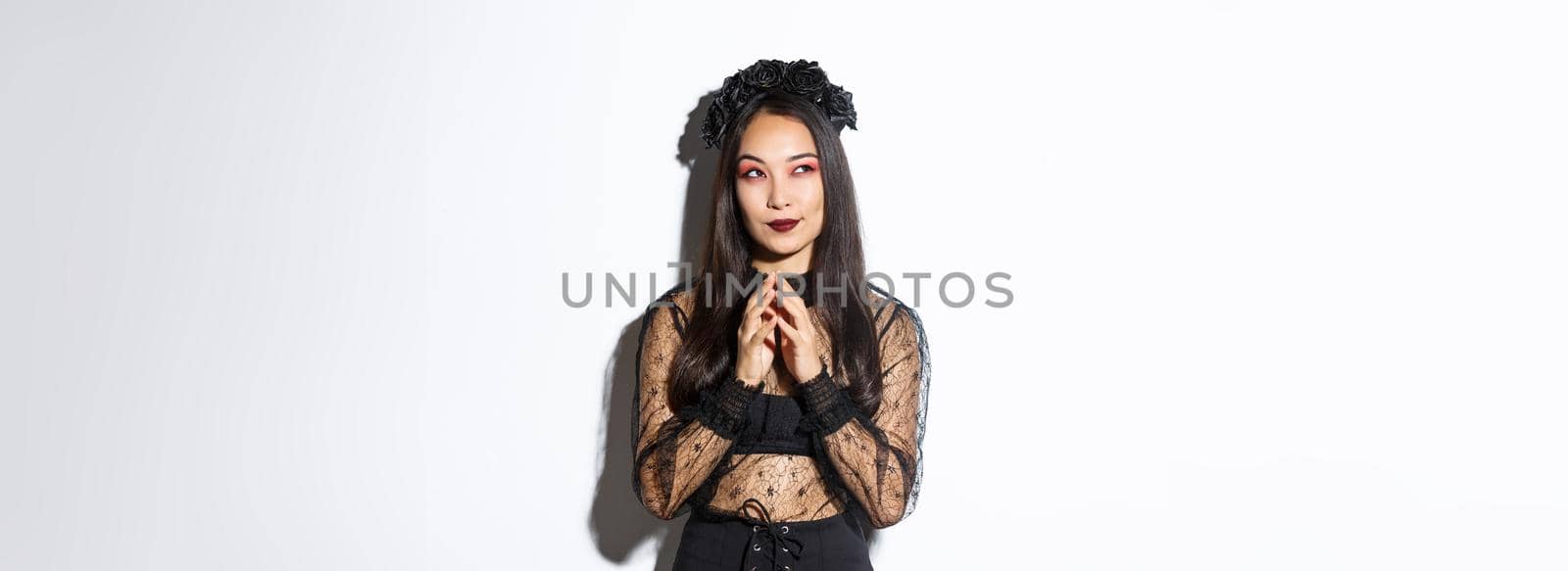 Thoughtful and cunning asian woman in witch costume prepared something for halloween, looking upper left corner and thinking. Girl wearing black gothic dress and wreath for party.