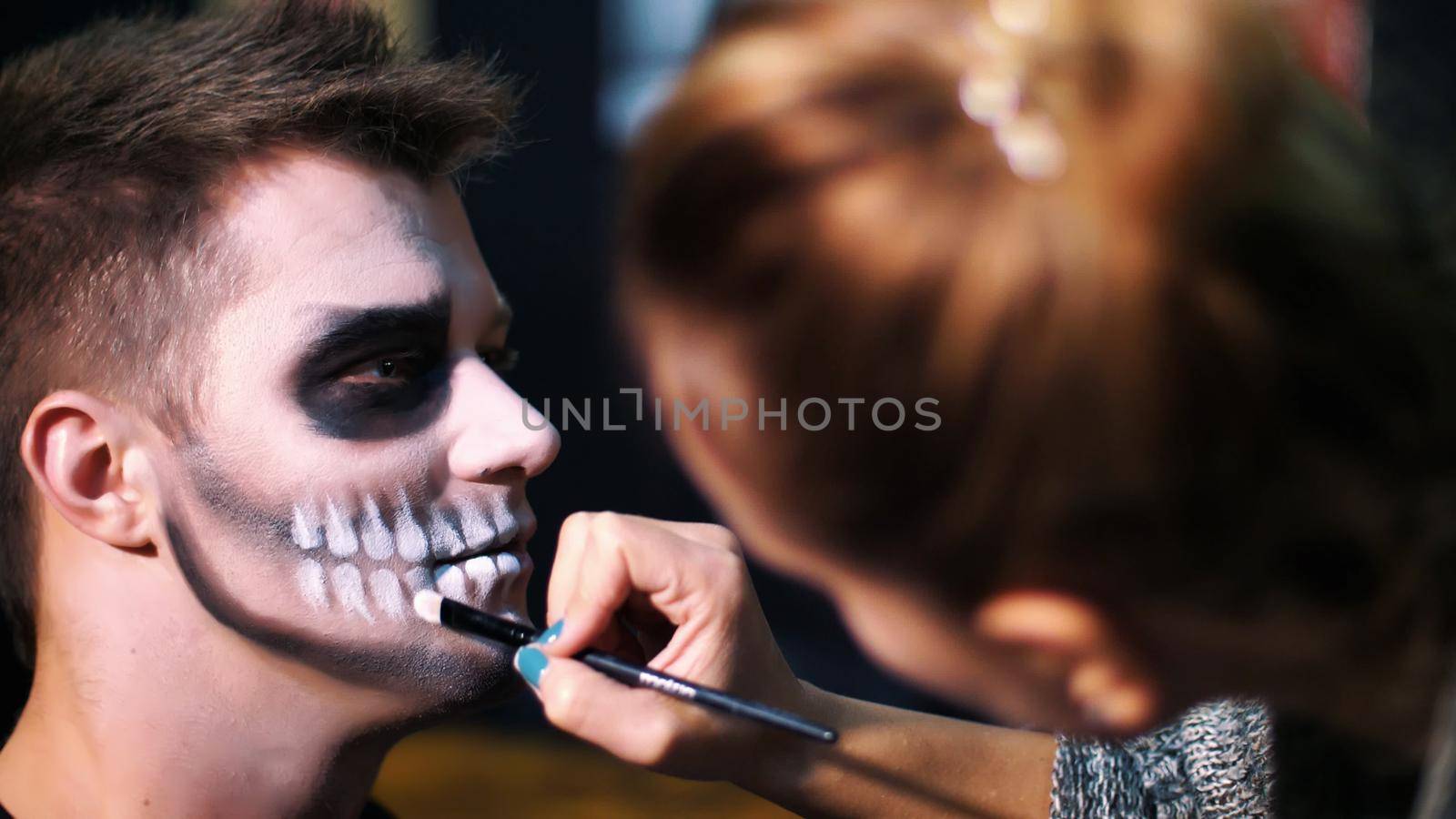 Halloween party, close-up, make-up artist draws a terrible makeup on the face of a man for a Halloween party. in the background the scenery in the style of Halloween is seen. High quality photo