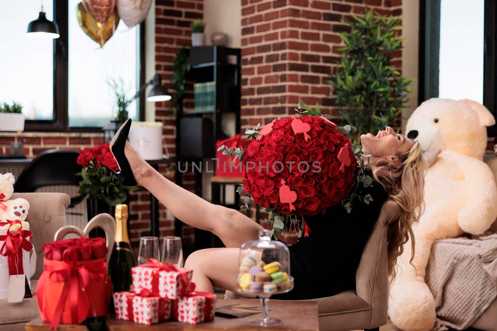 Woman holding red roses, laughing in room filled with luxury presents, romantic date. Excited beautiful blonde girl with expensive flower bouquet sitting, raising leg, side view