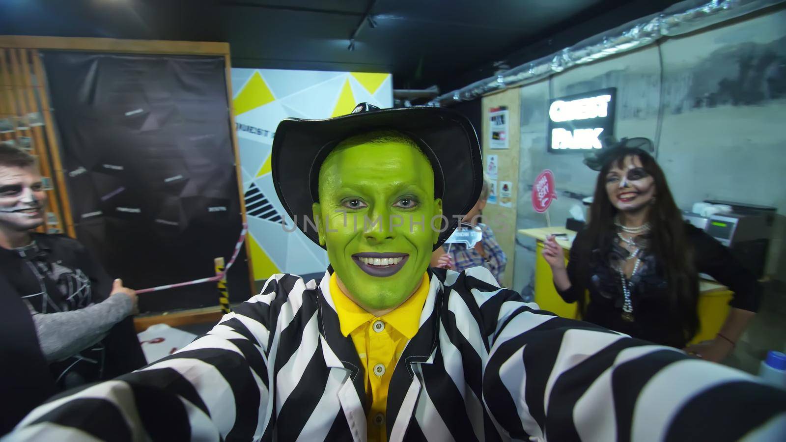 Halloween party, night, portrait of a man with a green face, wearing a hat, with a terrible makeup , croaks in front of the camera, in the background Halloween scenery is seen. High quality photo