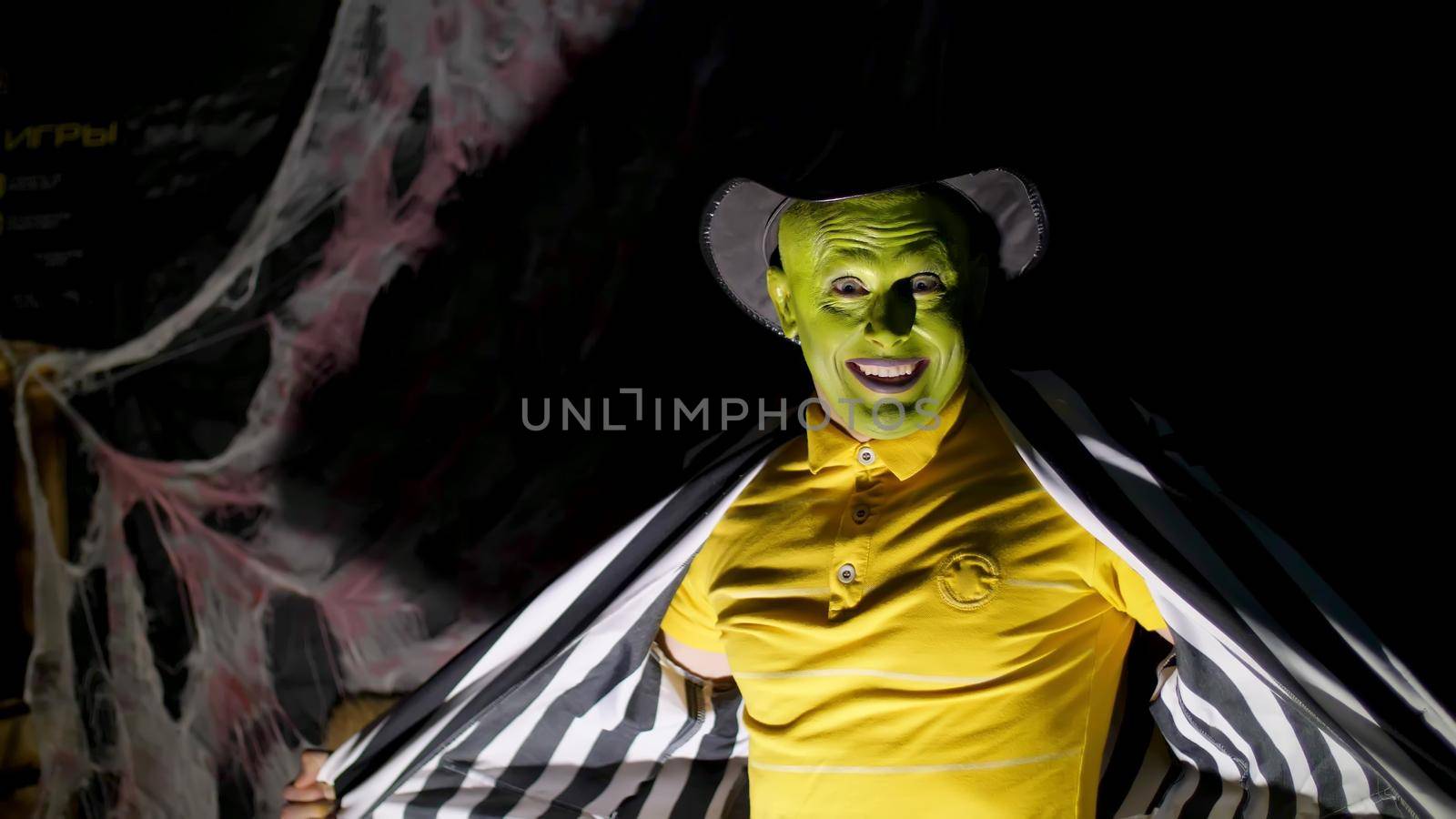 Halloween party, night, twilight, in the rays of light, a man with a terrible make-up, with a green face and a hat laughs eerily, frightens, Behind the background is a spiderweb. High quality photo