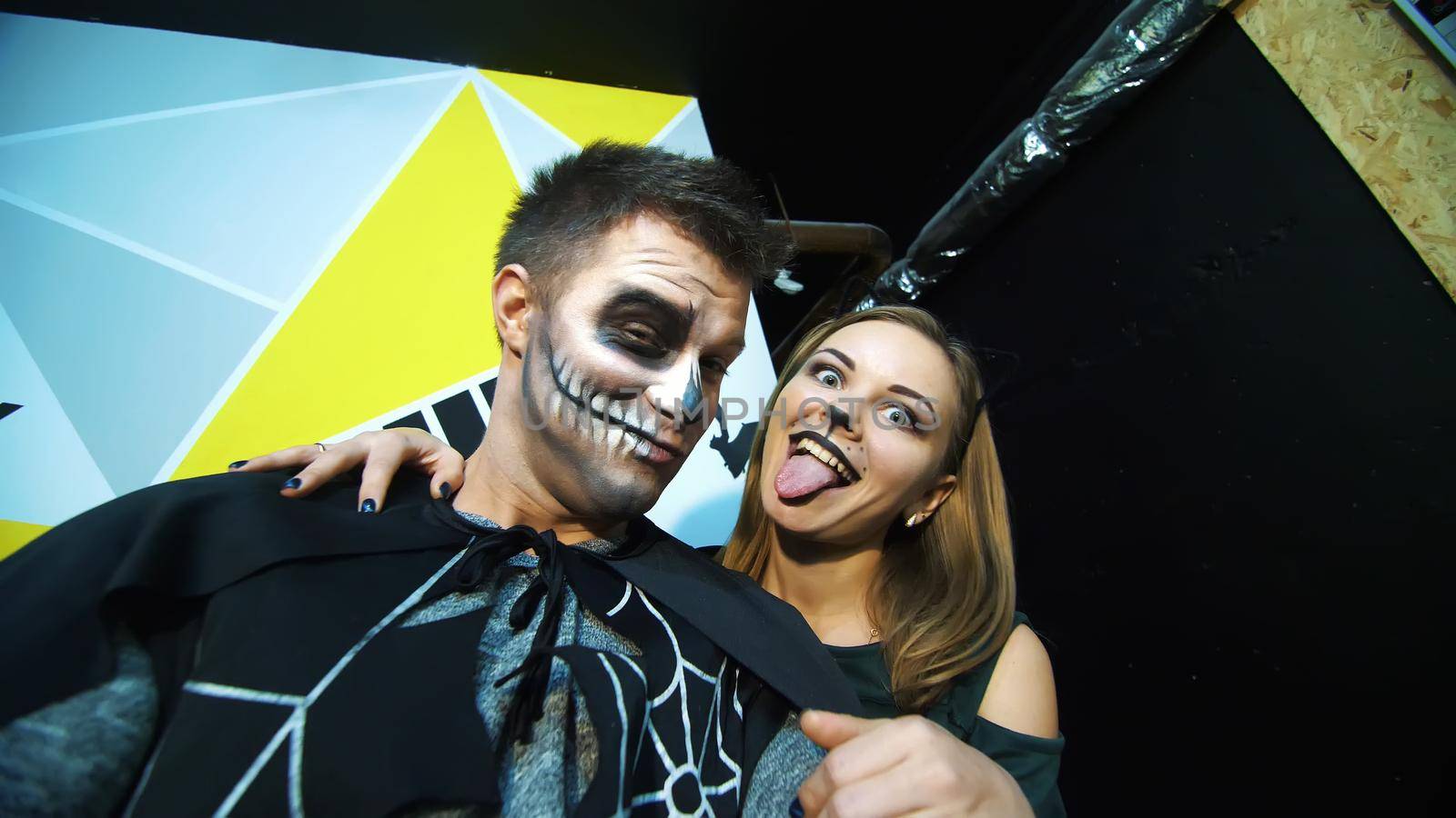 Halloween party, night, frightening portrait of a man with a terrible makeup in a black costume and a girl in a cat costume, they croak in front of the camera. High quality photo