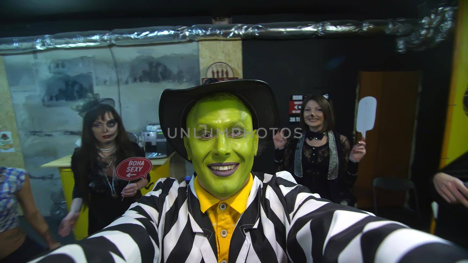 Halloween party, night, portrait of a man with a green face, wearing a hat, with a terrible makeup , croaks in front of the camera, in the background Halloween scenery is seen. High quality photo