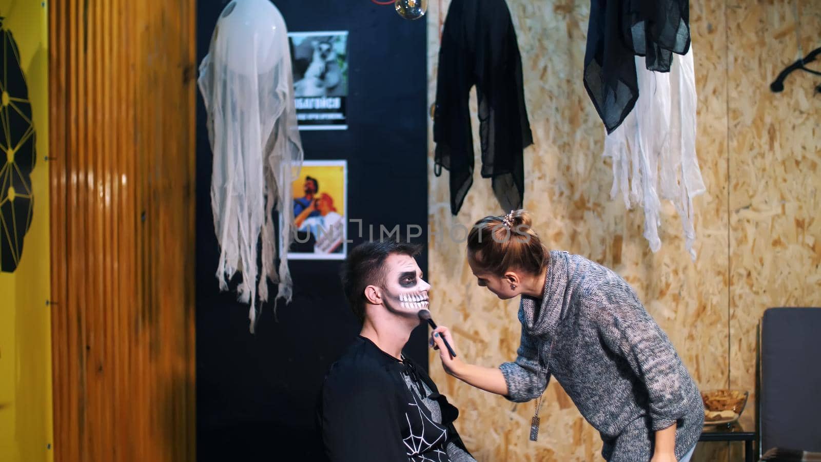 Halloween party, make-up artist draws a terrible makeup on the face of a man for a Halloween party. in the background, the scenery in the style of Halloween is seen by djtreneryay