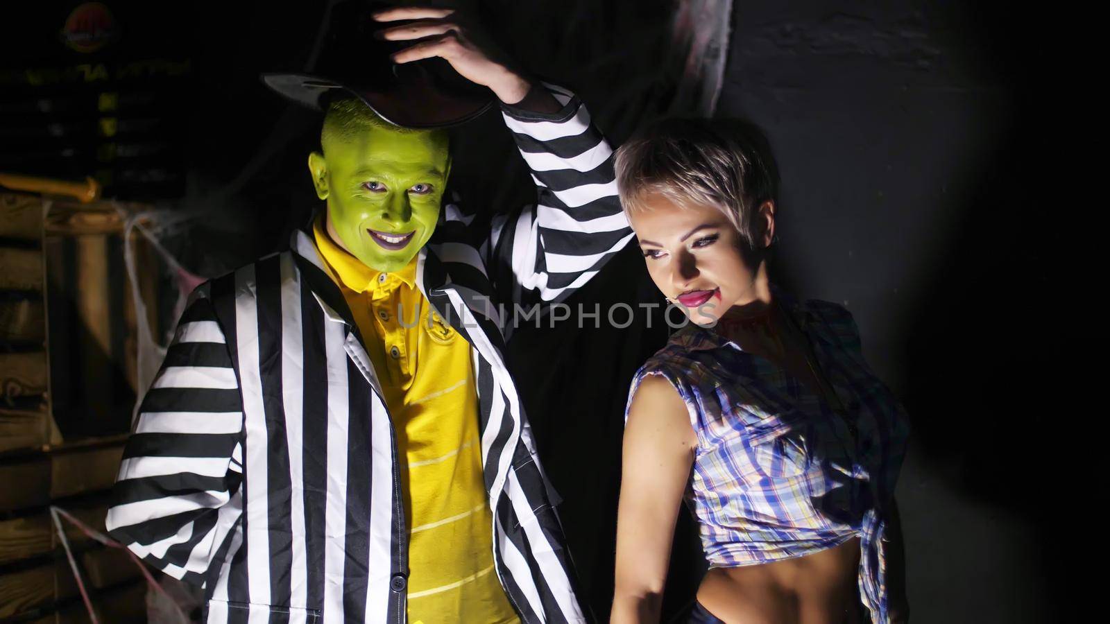 Halloween party, night, in the rays of light, a man with a terrible make-up, with a green face and a hat laughing eerily, pestering the girl in the schoolgirl's suit, slapping her ass, by djtreneryay