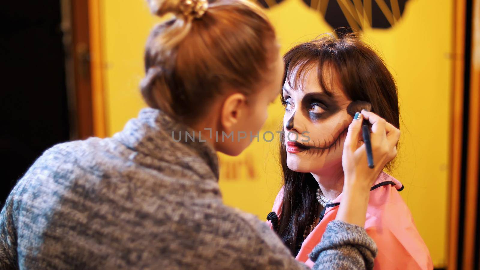 Halloween party, make-up artist draws a terrible makeup on the face of a brunette woman for a Halloween party. in the background, the scenery in the style of Halloween is seen. High quality photo