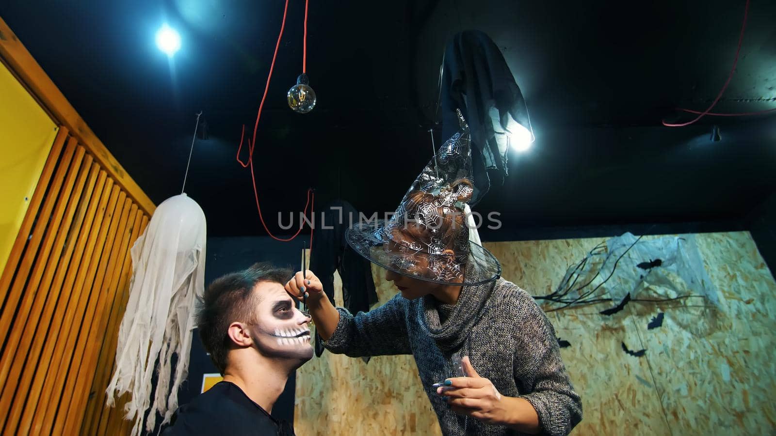 Halloween party, make-up artist draws a terrible makeup on the face of a man for a Halloween party. in the background, the scenery in the style of Halloween is seen. High quality photo