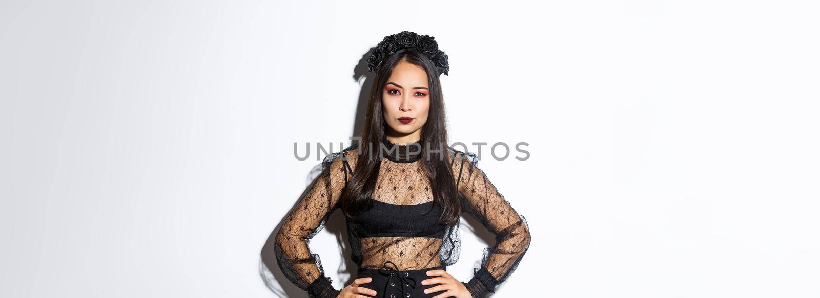 Attractive asian woman in halloween costume looking disappointed and skeptical. Female in black lace dress and wreath looking arrogant, trick or treat in witch outfit, standing white background.