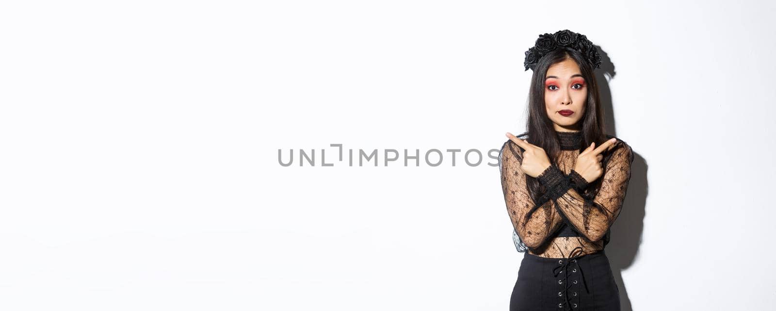 Confused pretty young woman in elegant black lace dress and wreath looking unsure, pointing fingers sideways, showing two variants, halloween banners, standing over white background.