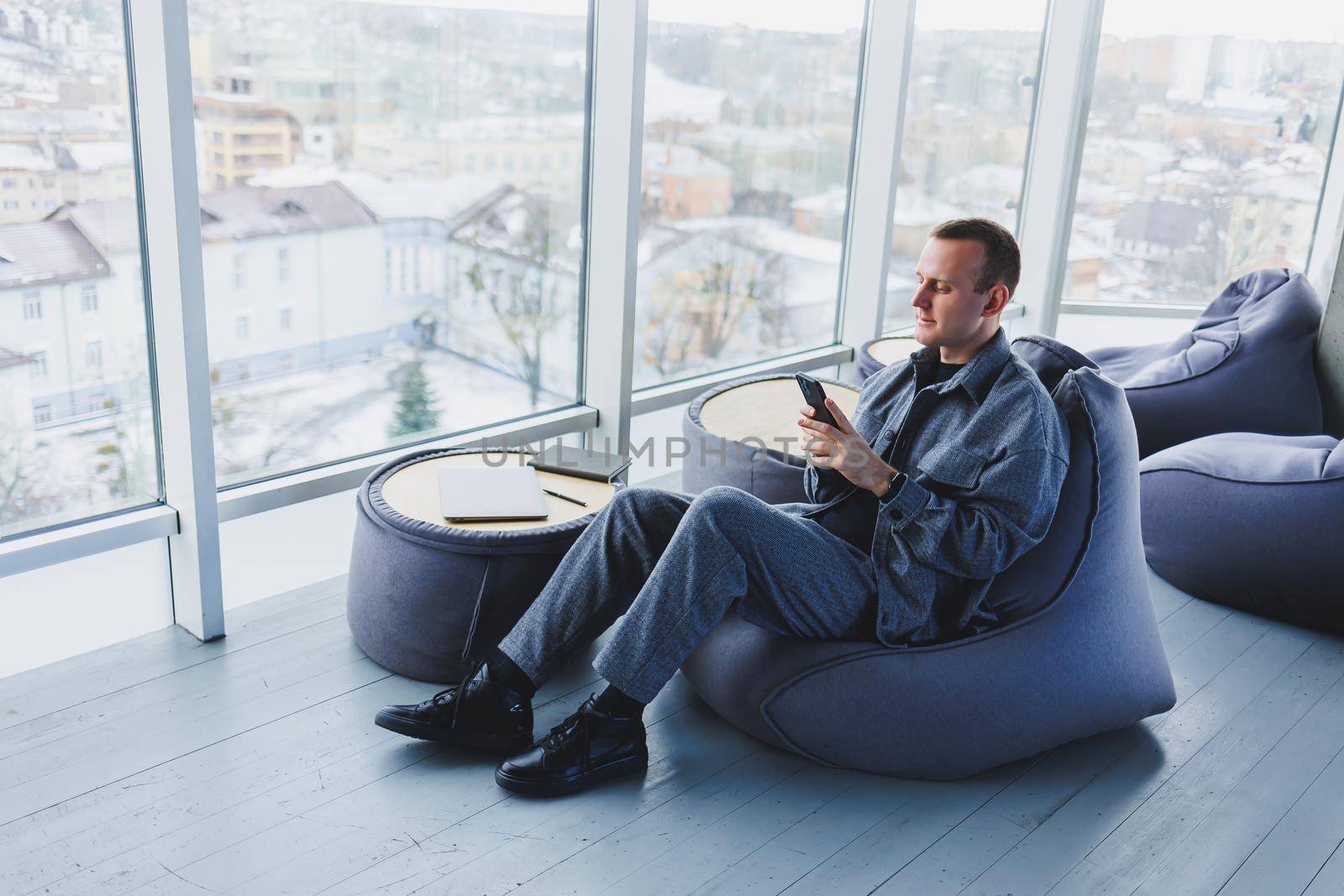 Young male student in a casual suit resting in a comfortable pouf while studying, an attractive freelancer in a suit works remotely
