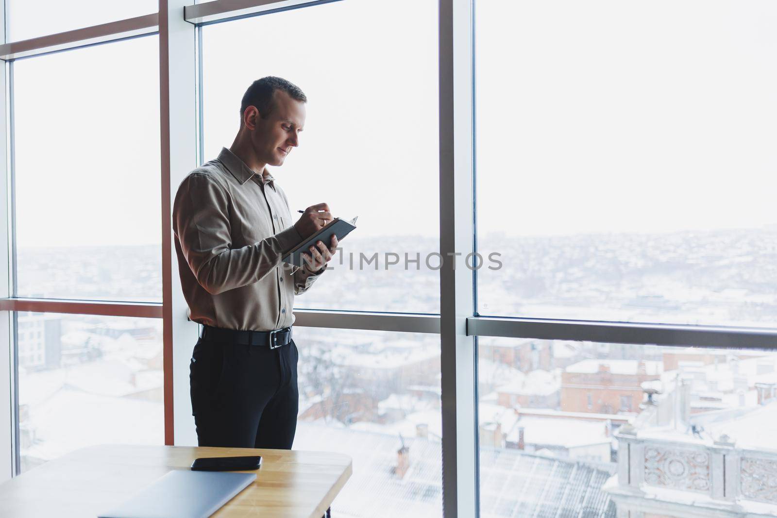 A young businessman stands at a panoramic window in a skyscraper and makes notes in his notebook with a pen. A man in a shirt and trousers of European appearance