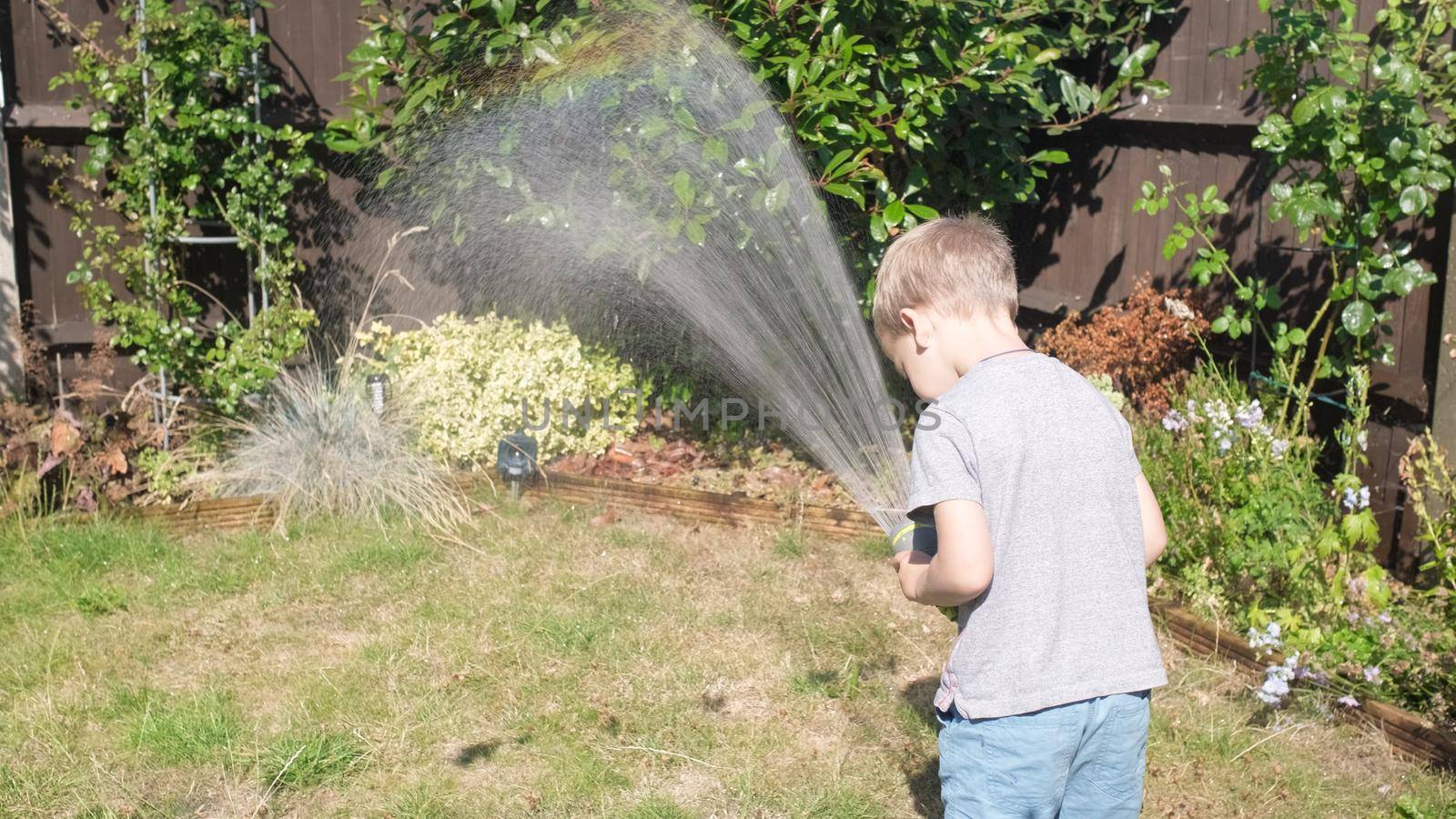Funny little boy watering lawn plants in garden housing backyard. Adorable child playing with irrigation hose at hot sunny summer outdoors. Children help with housework. activity for kids. Childhood.
