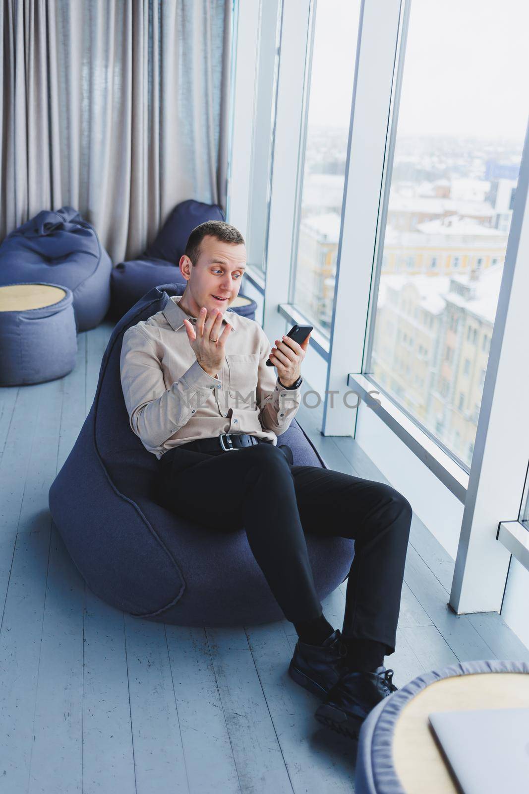 Successful male businessman sitting in a modern workspace and checking notifications on his mobile phone, he is wearing a shirt and trousers. Remote work.