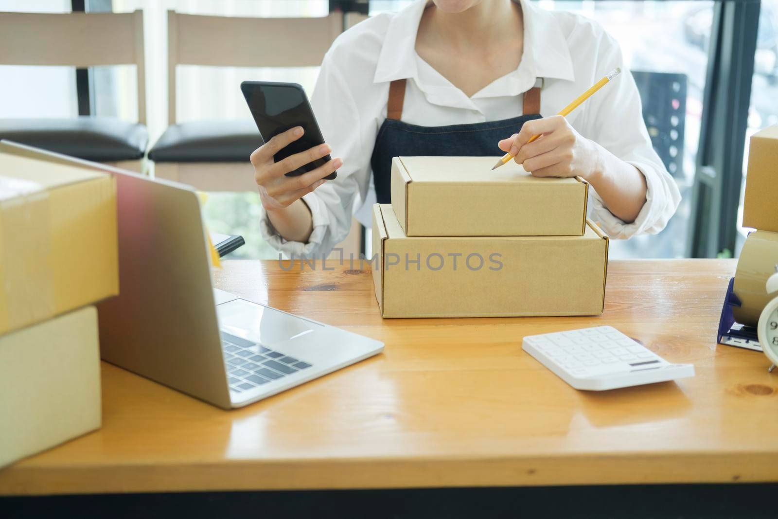 Asian female small online business or store owner packing shipping box and checking website retail order using cellphone preparing for delivery. Online business and e-coommerce concept.