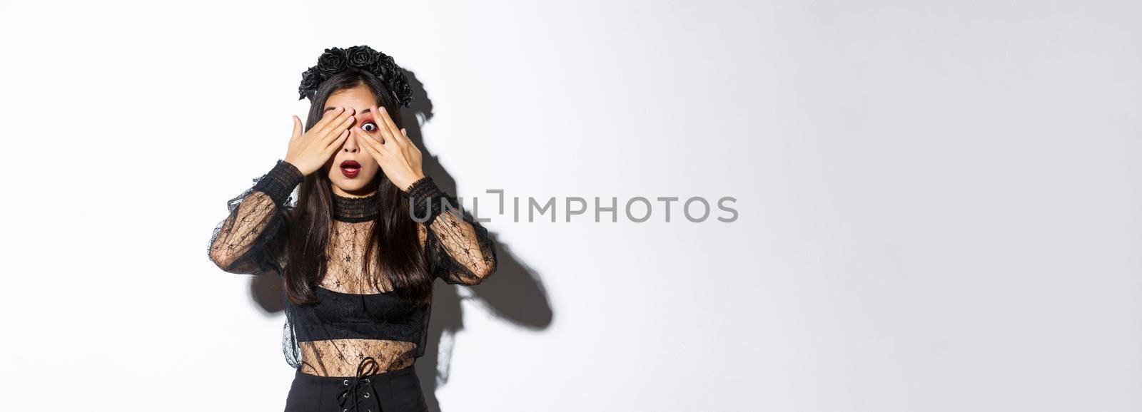 Image of attractive asian girl in halloween costume peeking at something curious, close eyes with fingers but looking through fingers, standing over white background by Benzoix
