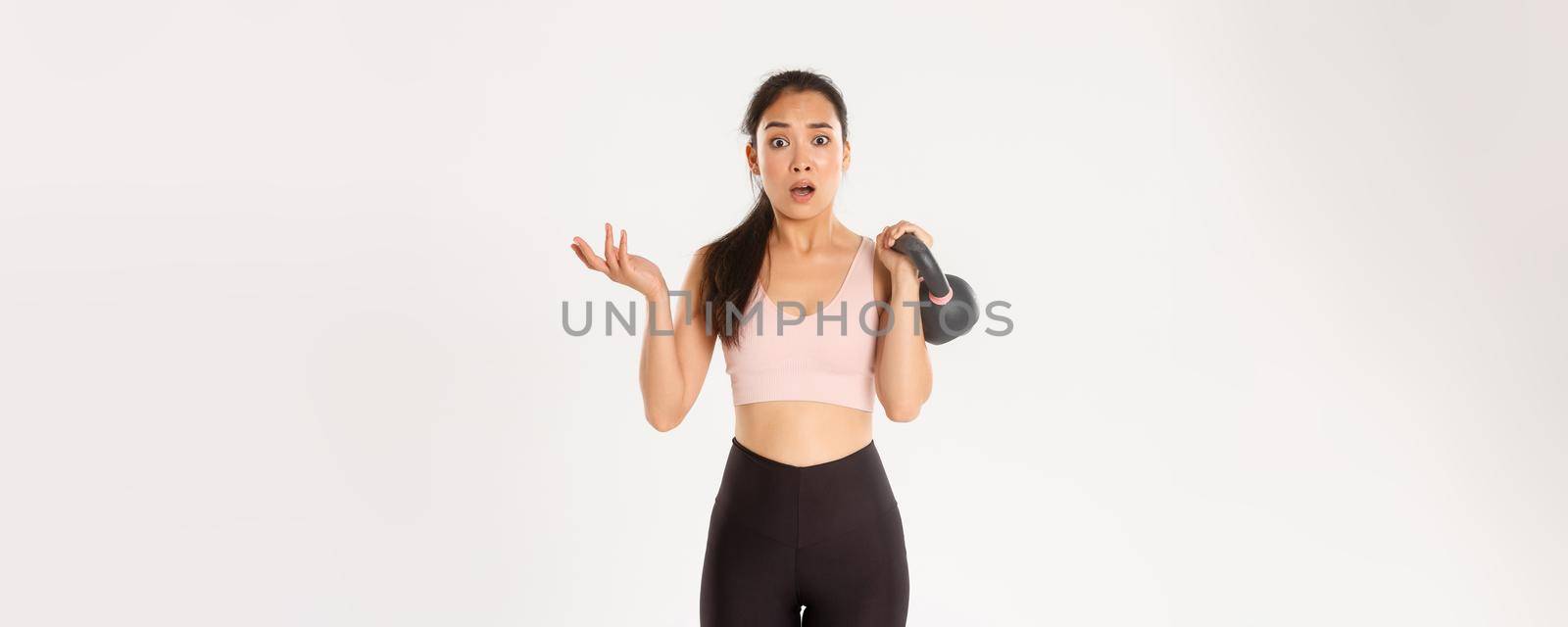 Sport, wellbeing and active lifestyle concept. Confused asian fitness girl, female athlete lifting kettlebell and look puzzled, consulting coach during training session, standing white background.