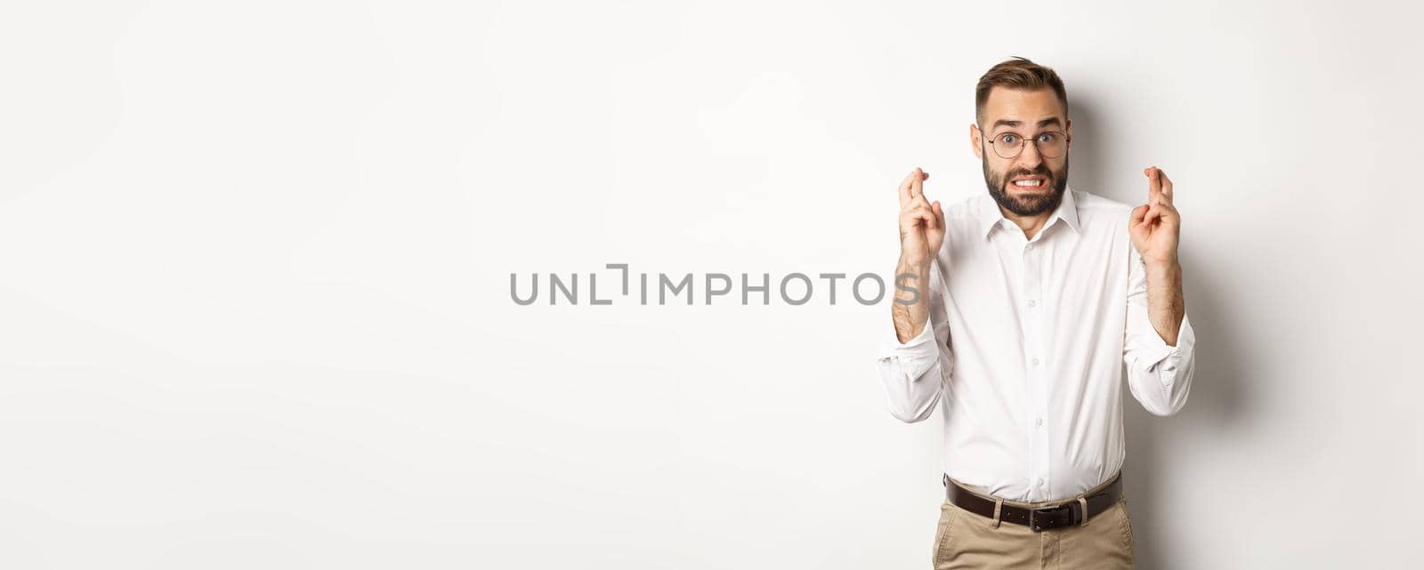 Worried man making a wish, cross fingers and hope for relish, standing over white background by Benzoix