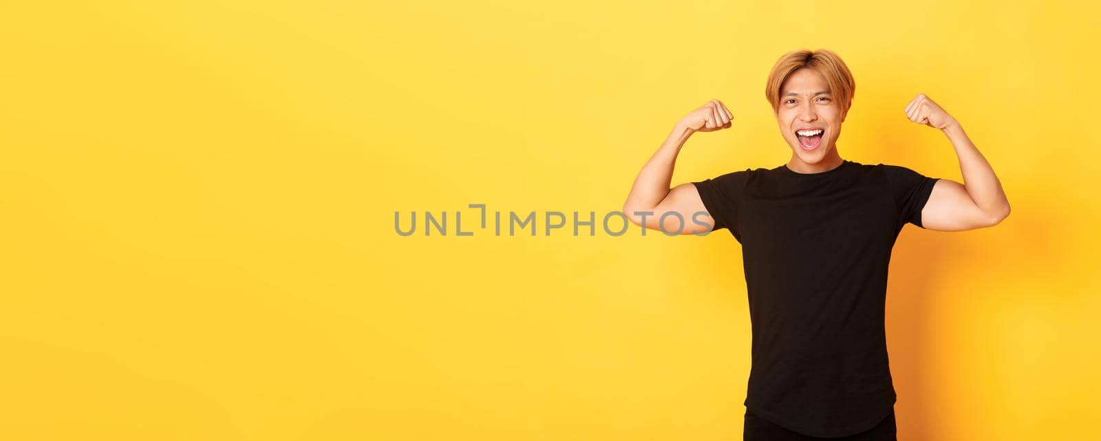 Portrait of happy and confident asian blond guy, flexing biceps, showing strong big muscles, standing yellow background.