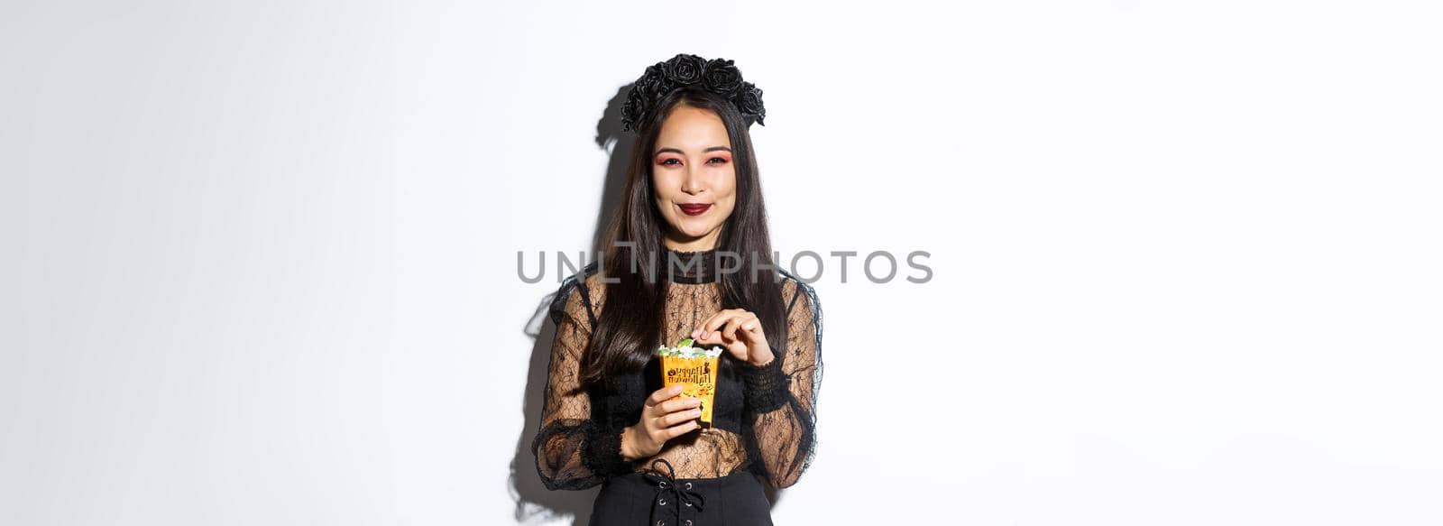 Smiling cute asian woman celebrating halloween, holding sweets and grinning happy, trick or treating in witch costume by Benzoix