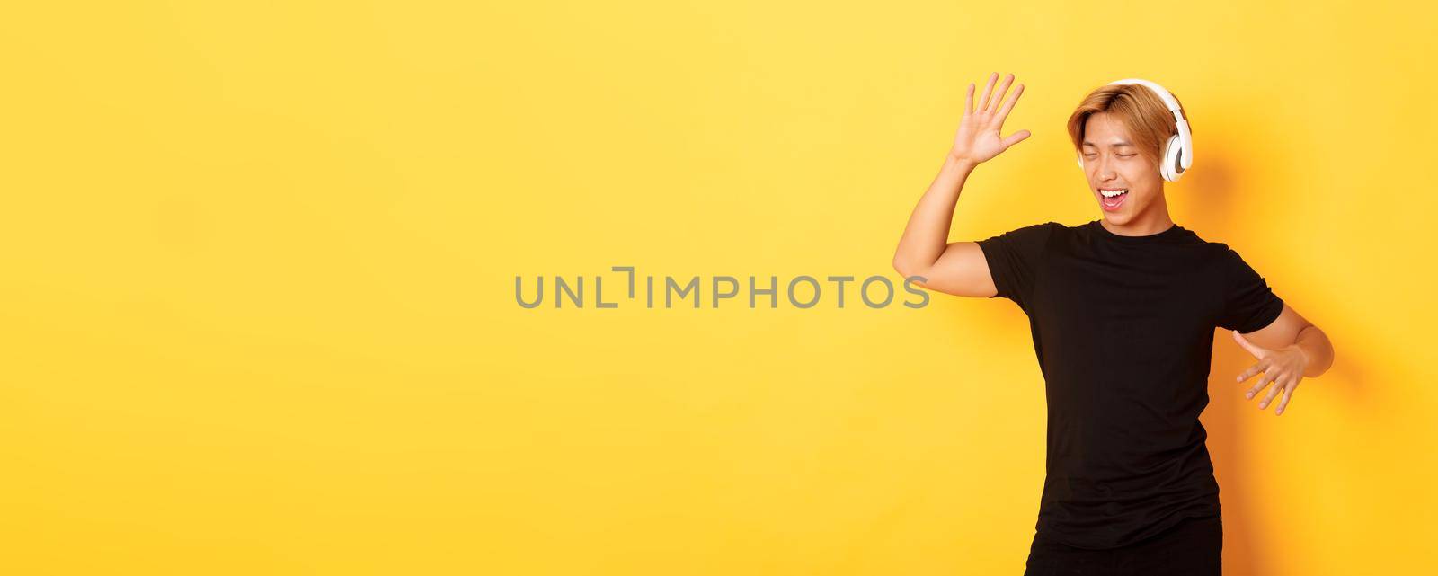 Happy attractive asian guy with blond hair, singing along and dancing as listening to music in wireless headphones, standing yellow background by Benzoix