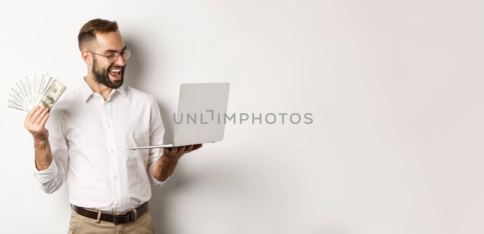 Business and e-commerce. Satisfied businessman doing job on laptop and holding money, smiling happy, standing over white background.