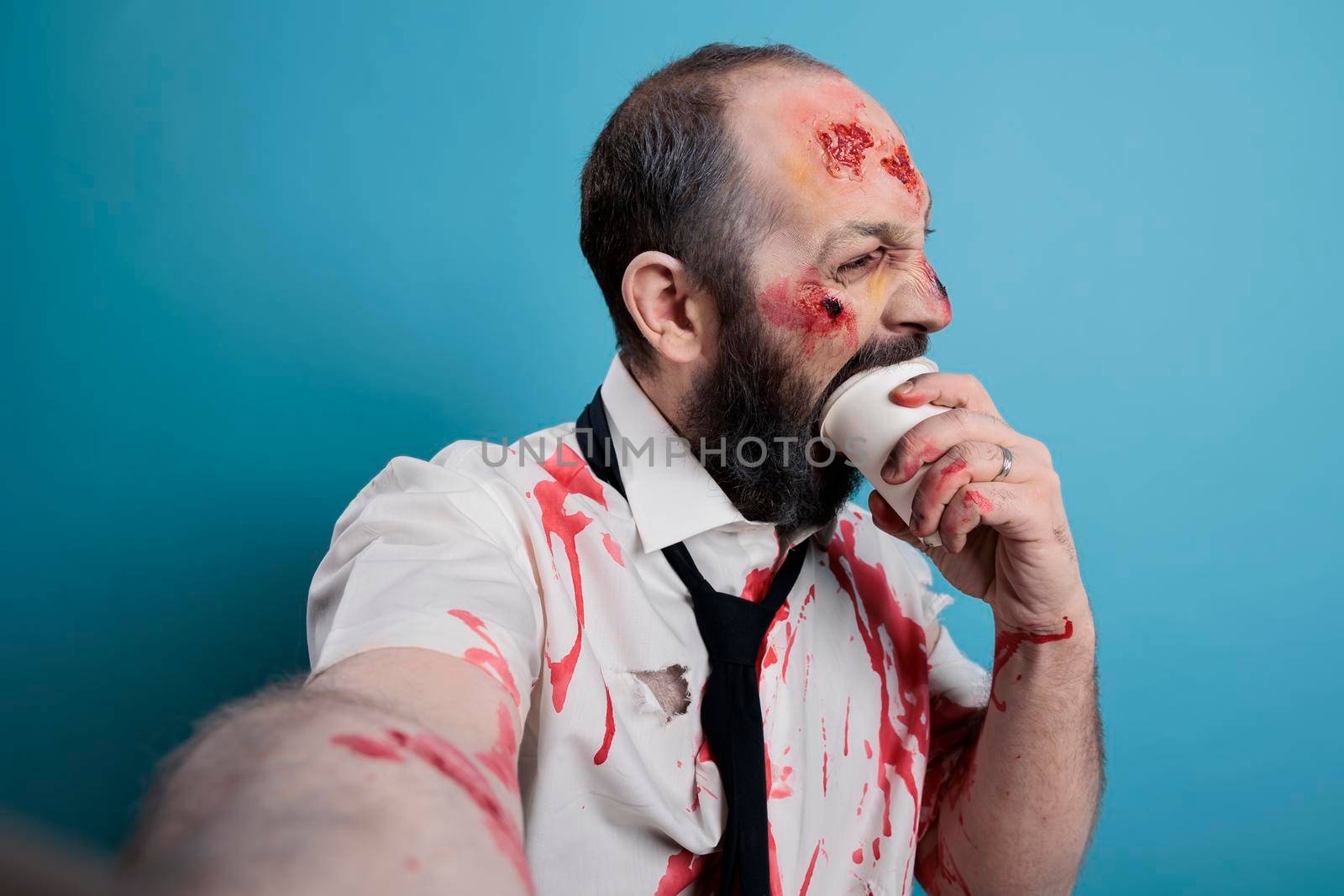 Dangerous halloween zombie biting on coffee cup and acting aggressive, looking creepy and taking photo in studio. Undead evil monster corpse with bloody scars eating carton, doomsday.