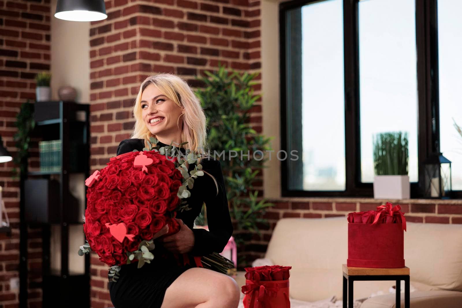 Woman posing with luxury red roses bouquet, laughing, sitting on bar stool in living room, medium shot. Beautiful blonde girl with valentines day expensive gift, romantic date.