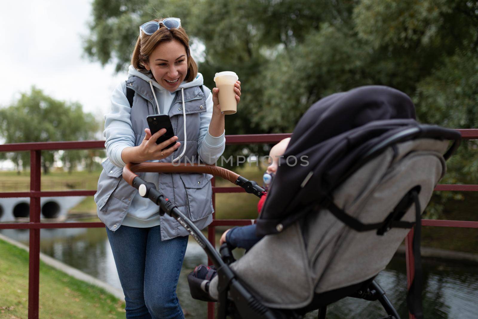 a young mother sipping coffee in headphones walks in the park with a baby stroller.