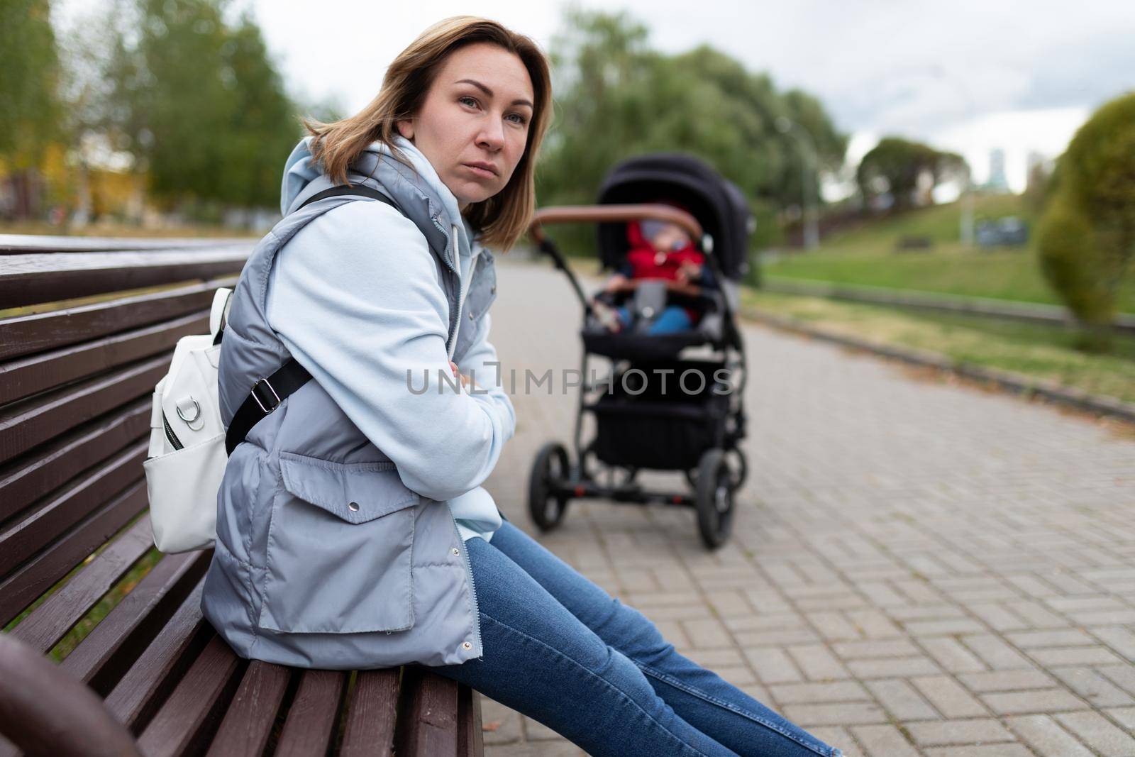 annoyed young mother sitting in the park on a bench away from the baby stroller with an angry face. the concept of postpartum depression.
