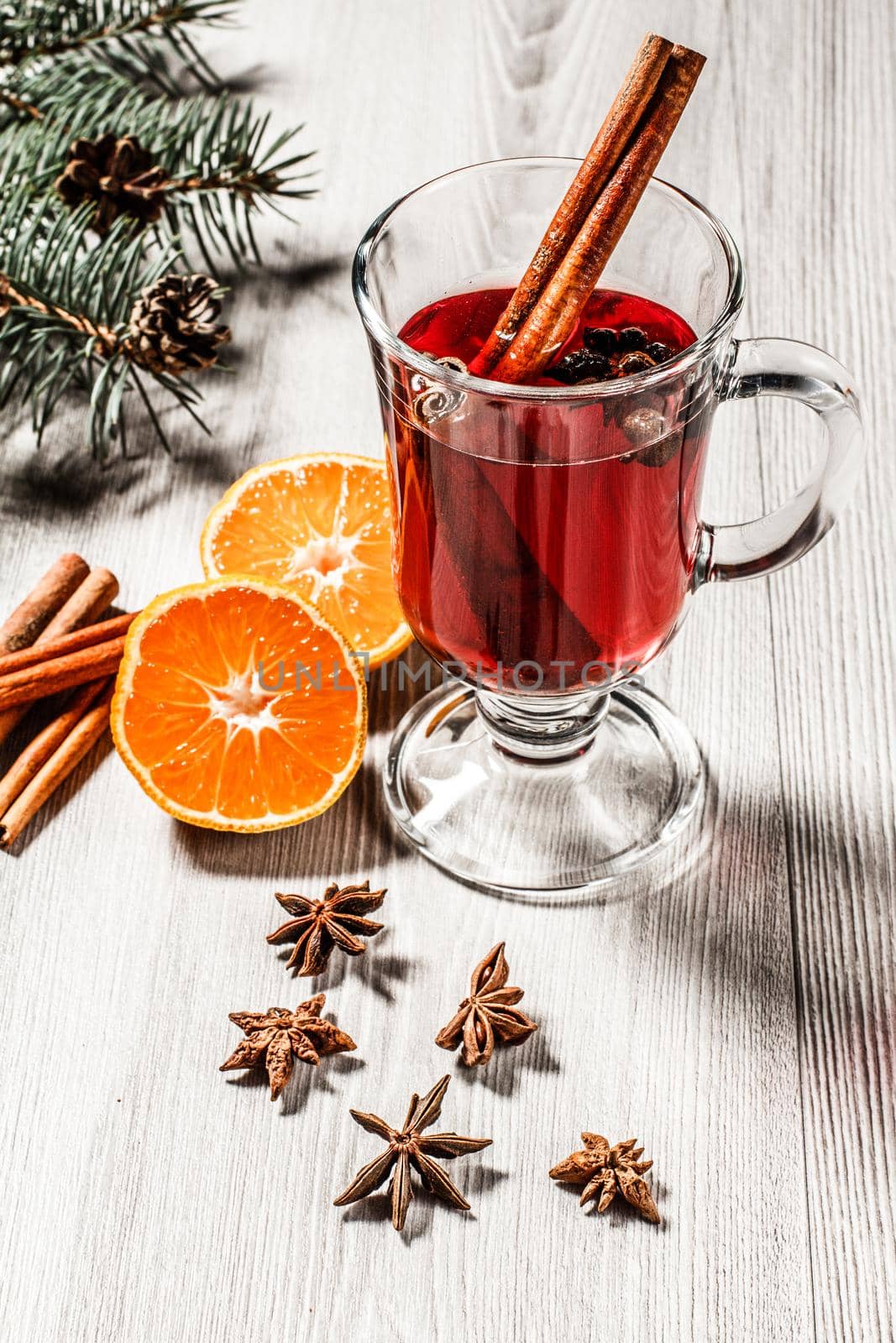 Glass of Christmas mulled wine with cinnamon, star anise and cloves on wooden background with white biscuits, slices of orange, natural fir tree branches and cones. Color toning effect.