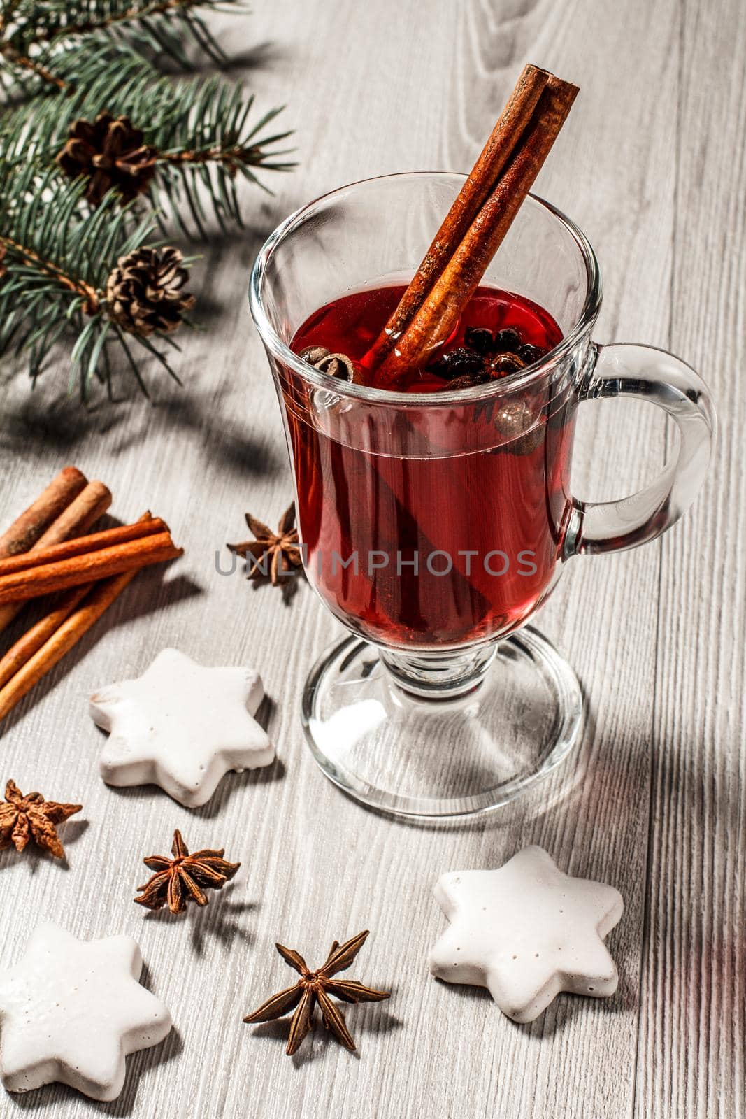 Glass of Christmas mulled wine with cinnamon, star anise and cloves on wooden background with white biscuits, natural fir tree branches and cones. Color toning effect.