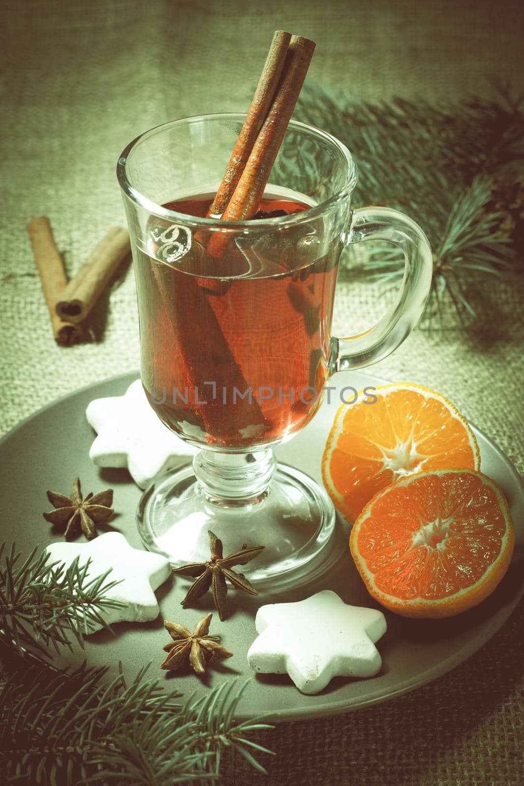Glass of Christmas mulled wine with cinnamon, star anise and cloves on plate with slices of orange, white biscuits and natural fir tree branches on the background. Color toning effect.