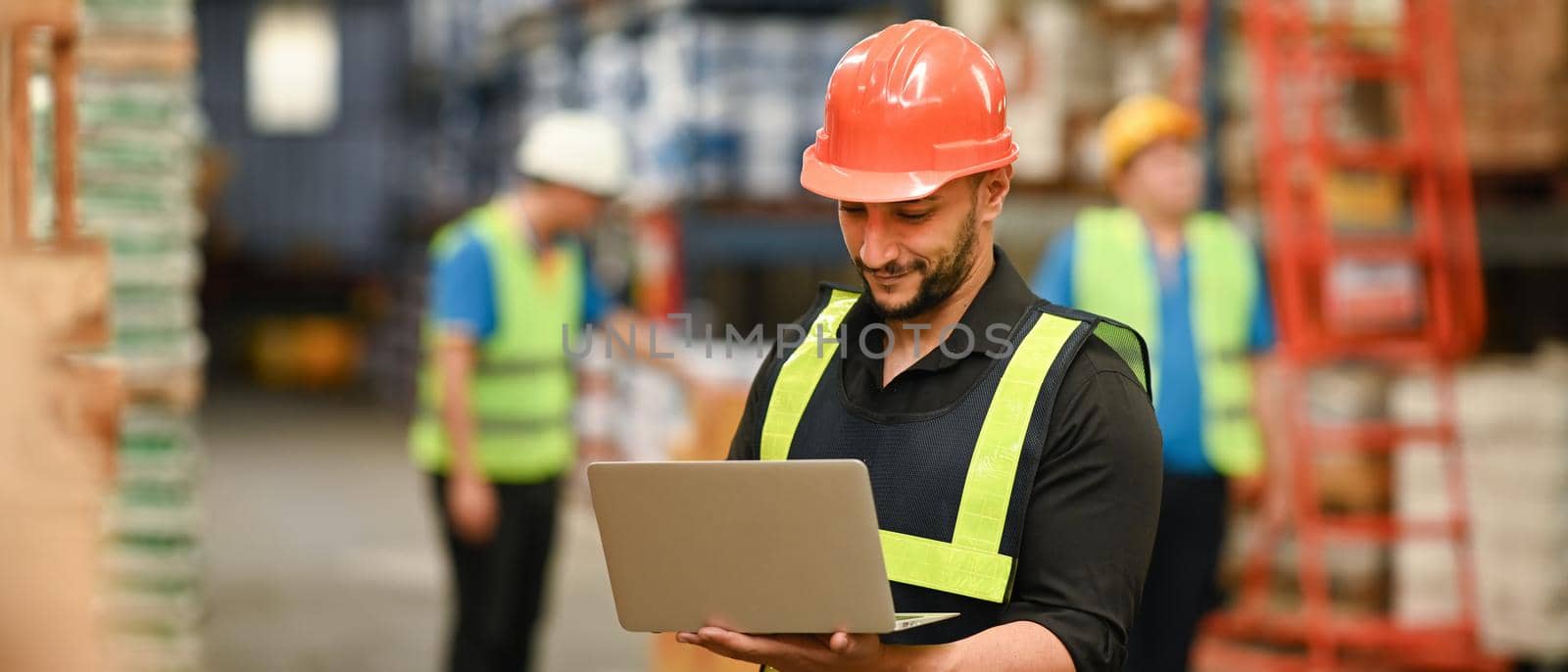 Handsome male warehouse manager in hardhats and reflective jackets on laptop for checking stock and order details by prathanchorruangsak