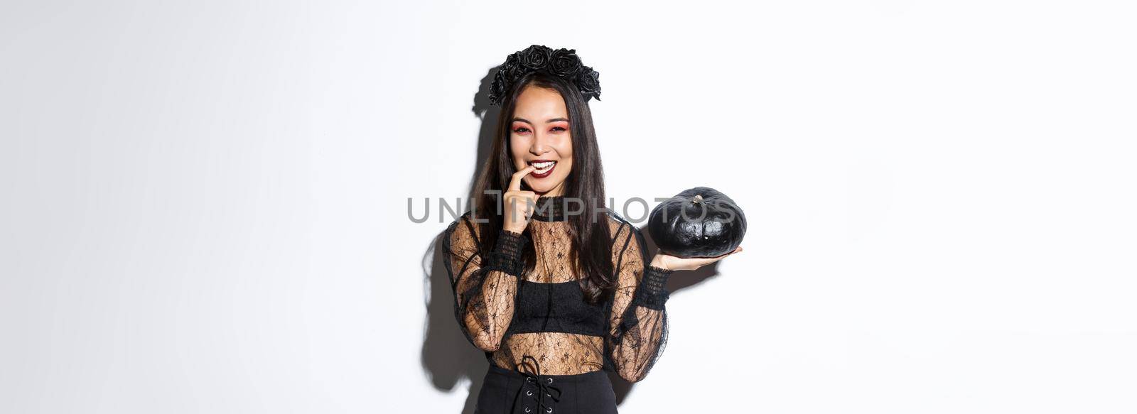 Sassy attractive asian woman in witch costume looking cunning and smiling, holding pumpking, going trick or treating on halloween, standing over white background.