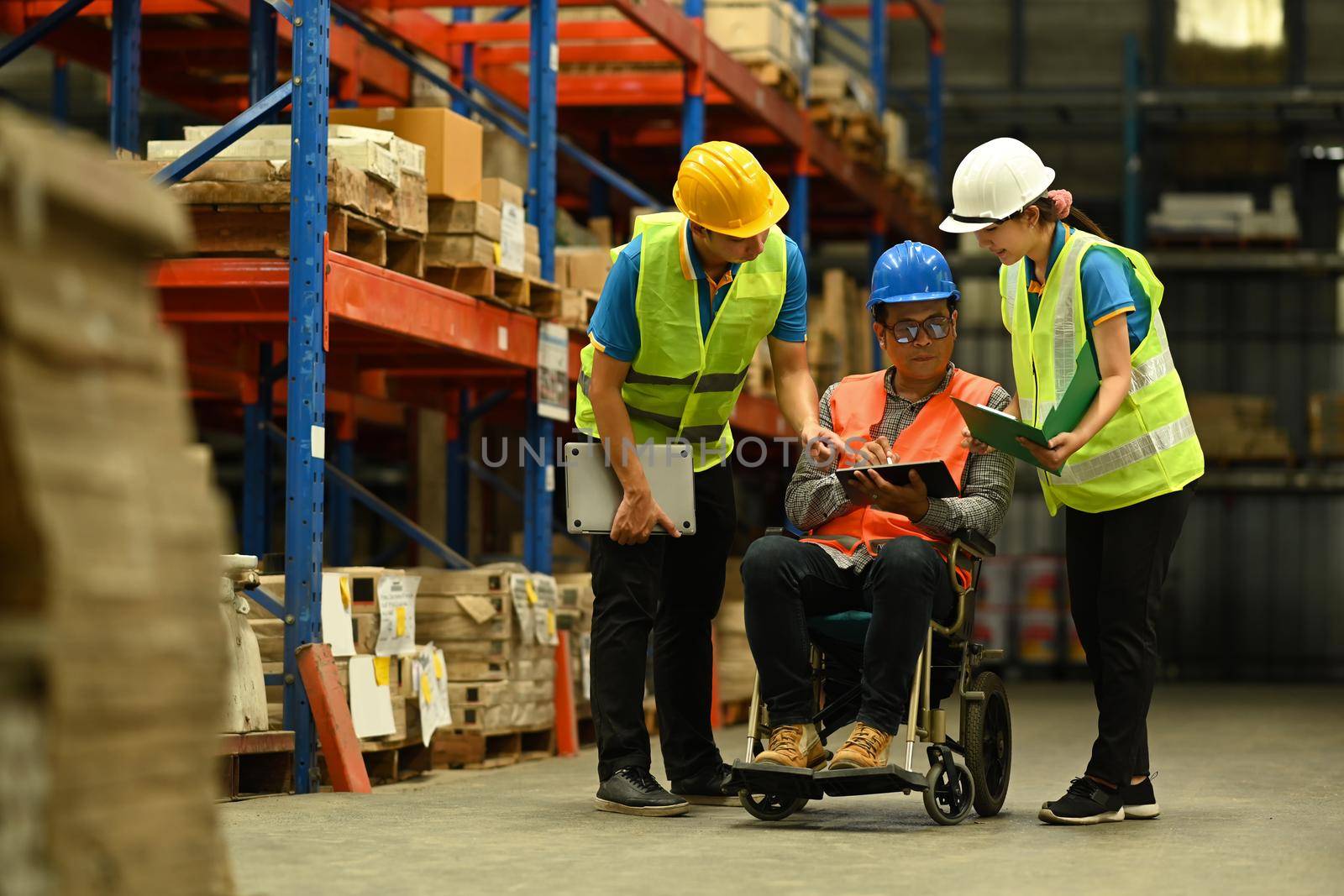 Asian male manager in wheelchair and young workers using digital tablet, inspecting stock product in a large warehouse.