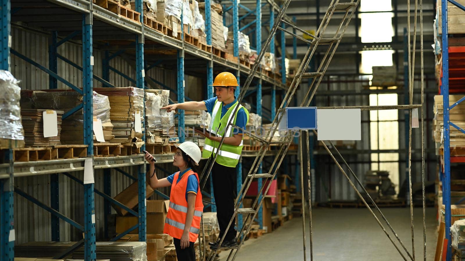 Male and female workers wearing hardhats and reflective jackets checking inventory boxes on shelf with barcode scanner.