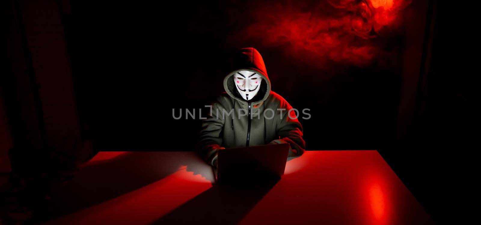 Anonymous in a hood is typing on a laptop in the dark in red smoke. by mrwed54