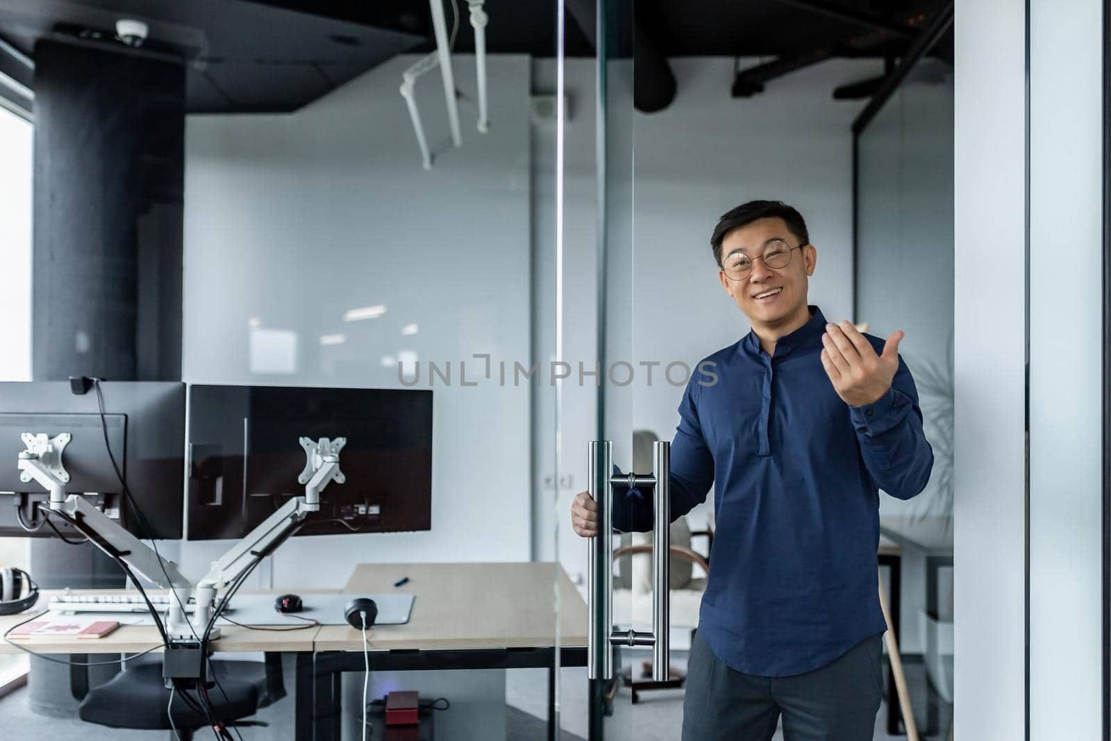 A successful engineer programmer boss invites to his office, an Asian man in a shirt and glasses looks at the camera and smiles and waves to the interlocutor.