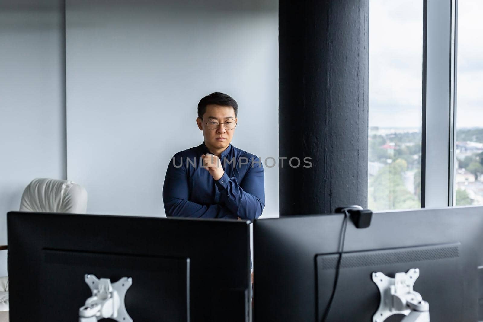 Serious and thinking asian engineer looking at two monitors, close-up photo of office worker, man working inside modern office building, businessman wearing glasses and casual shirt.