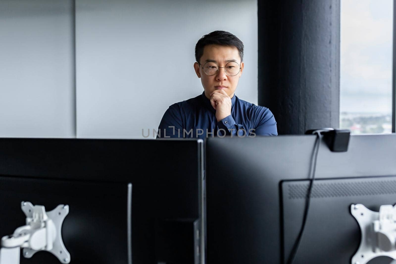 Serious and thinking asian engineer looking at two monitors, close-up photo of office worker, man working inside modern office building, businessman wearing glasses and casual shirt.
