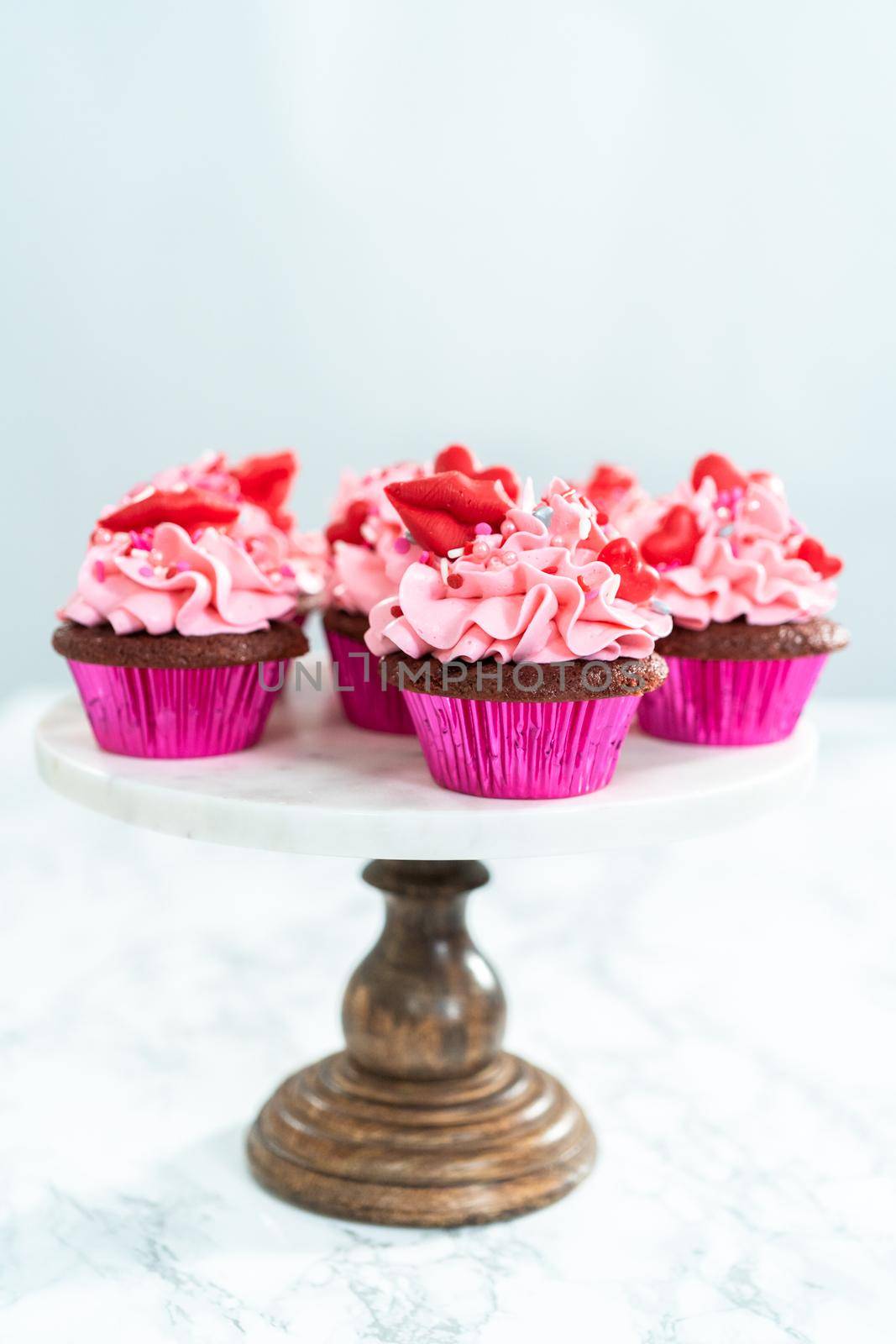 Red velvet cupcakes with pink Italian buttercream frosting and decorates with heart and kiss shaped red chocolates.