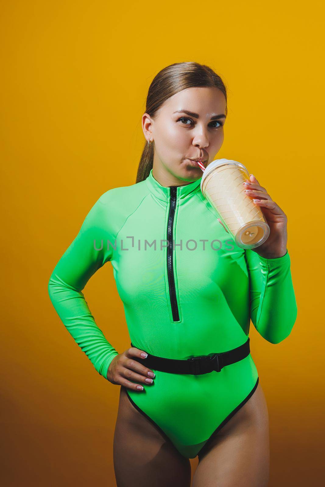 A young smiling blonde in a green swimsuit with a cocktail in her hands is happy about the upcoming vacation, isolated on bright yellow. Summer tropics travel concept.